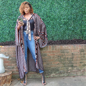 A woman models a flowing kimono jacket in black, grey, and white with a hood and a trim adorned with a horse and carriage motif, blending chic design with grace.